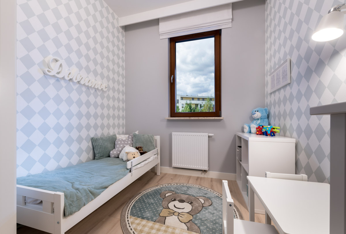 Ile trwa wykończenie domu pod klucz - zdjęcie przedstawia mały pokój dziecięcy z pojedynczym łóżkiem, dywanem z wizerunkiem pluszowego misia, biała szafka i mały stolik. Pomieszczenie jest utrzymane w kolorystyce biało - miętowej