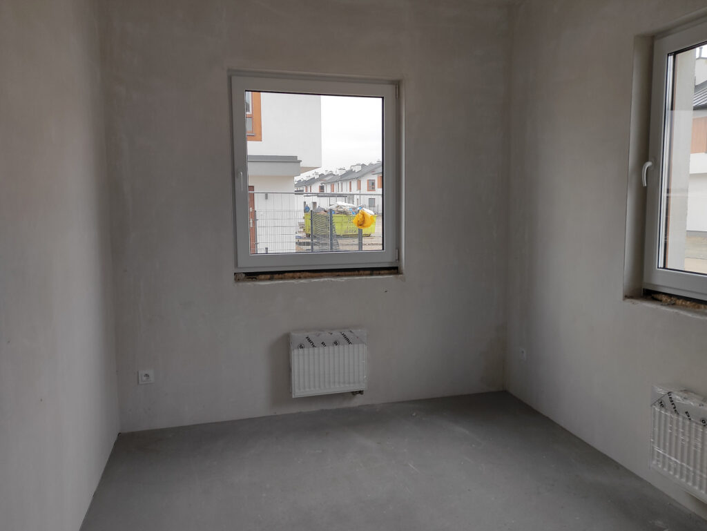 Wykończenie domu pod klucz - zdjęcie przedstawia małe pomieszczenie w stanie deweloperskim. W pomieszczeniu znajdują się dwa małe okna kaloryfery i puste ściany